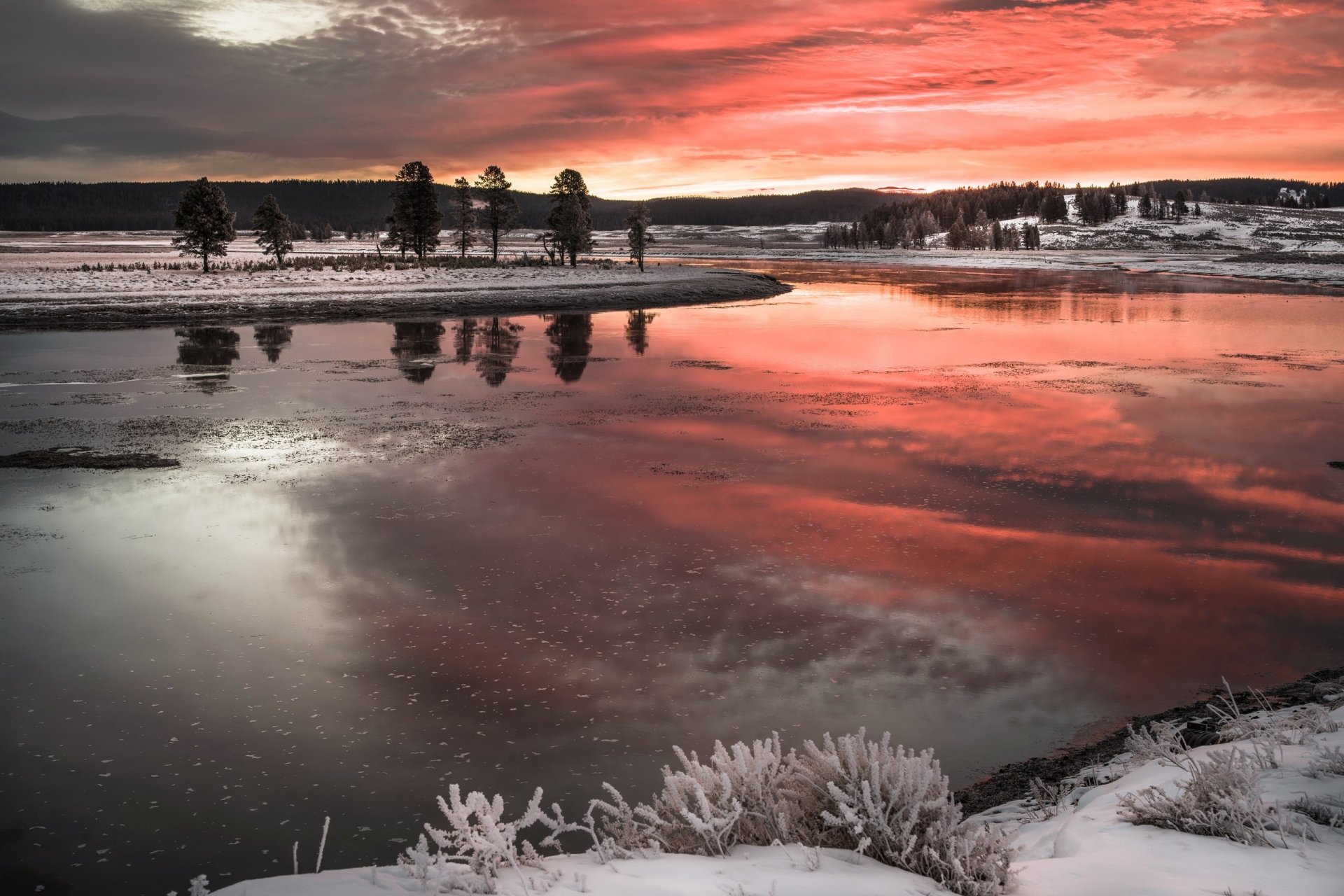 2K Quad HD PC desktop wallpaper: winter sunset over a lake in a quiet nature scene, trees along the shore and fiery orange-red sky reflected in the icy water, snowy foreground.