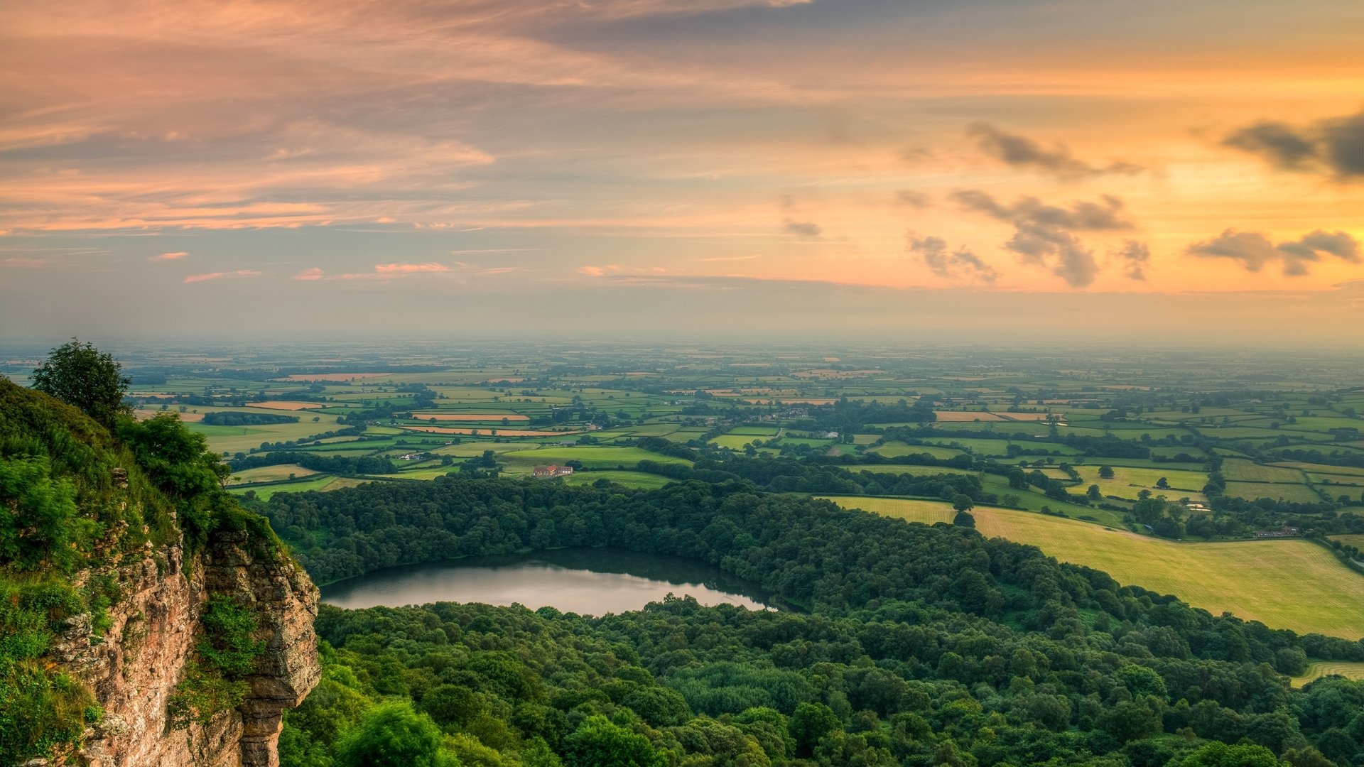 HD desktop wallpaper featuring a serene lake surrounded by lush green fields under a colorful sunset sky, capturing the beauty of nature and a distant horizon.
