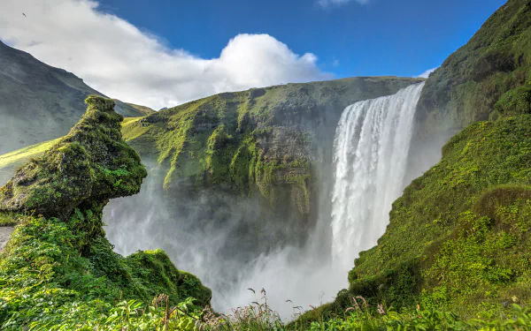 HD PC desktop wallpaper of Skógafoss waterfall in Iceland, water cascading between green mossy mountains beneath a bright blue sky — lush nature background.