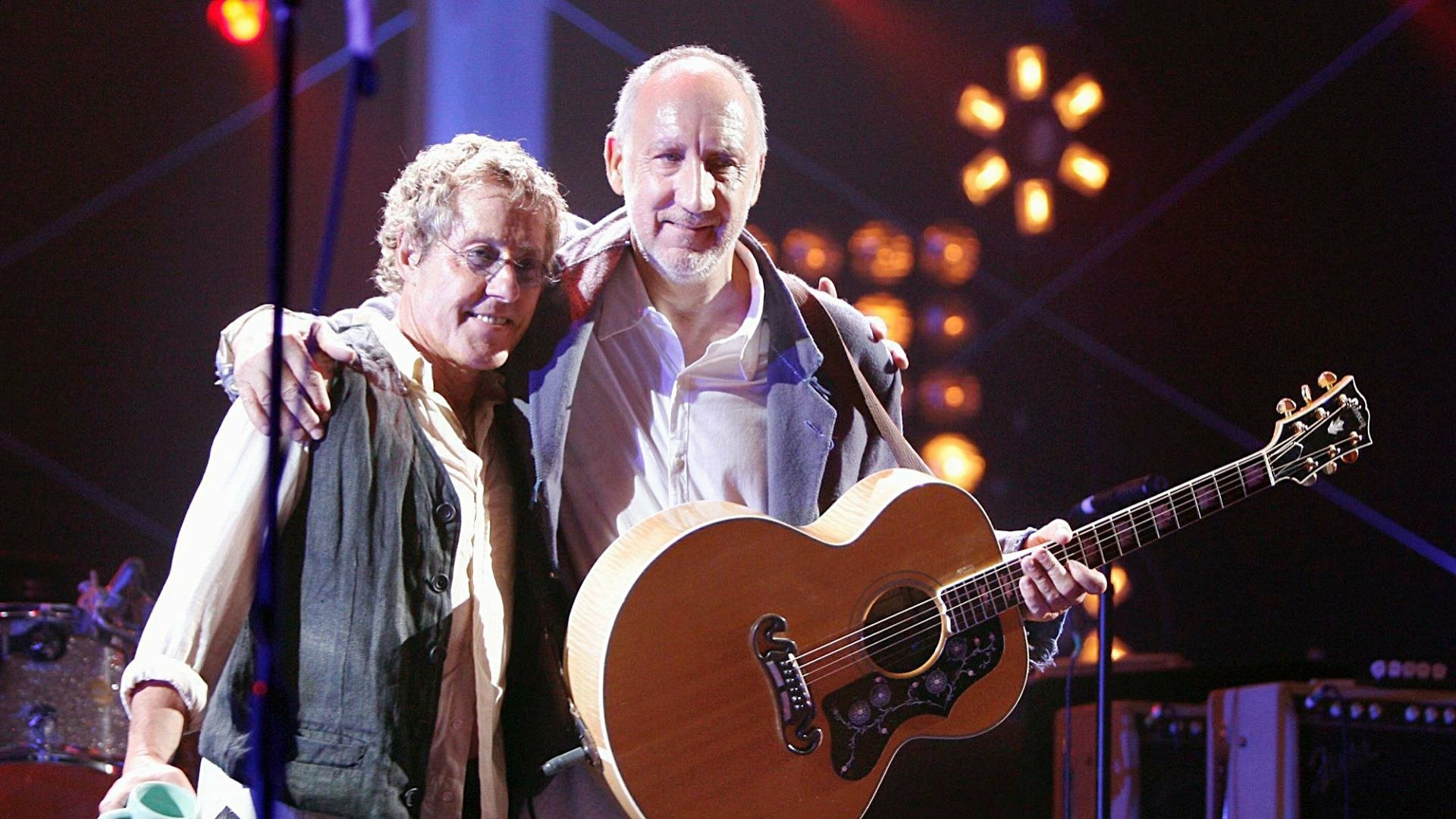HD PC desktop wallpaper showing two rock musicians onstage, one holding an acoustic guitar under warm stage lights, music-themed concert background.