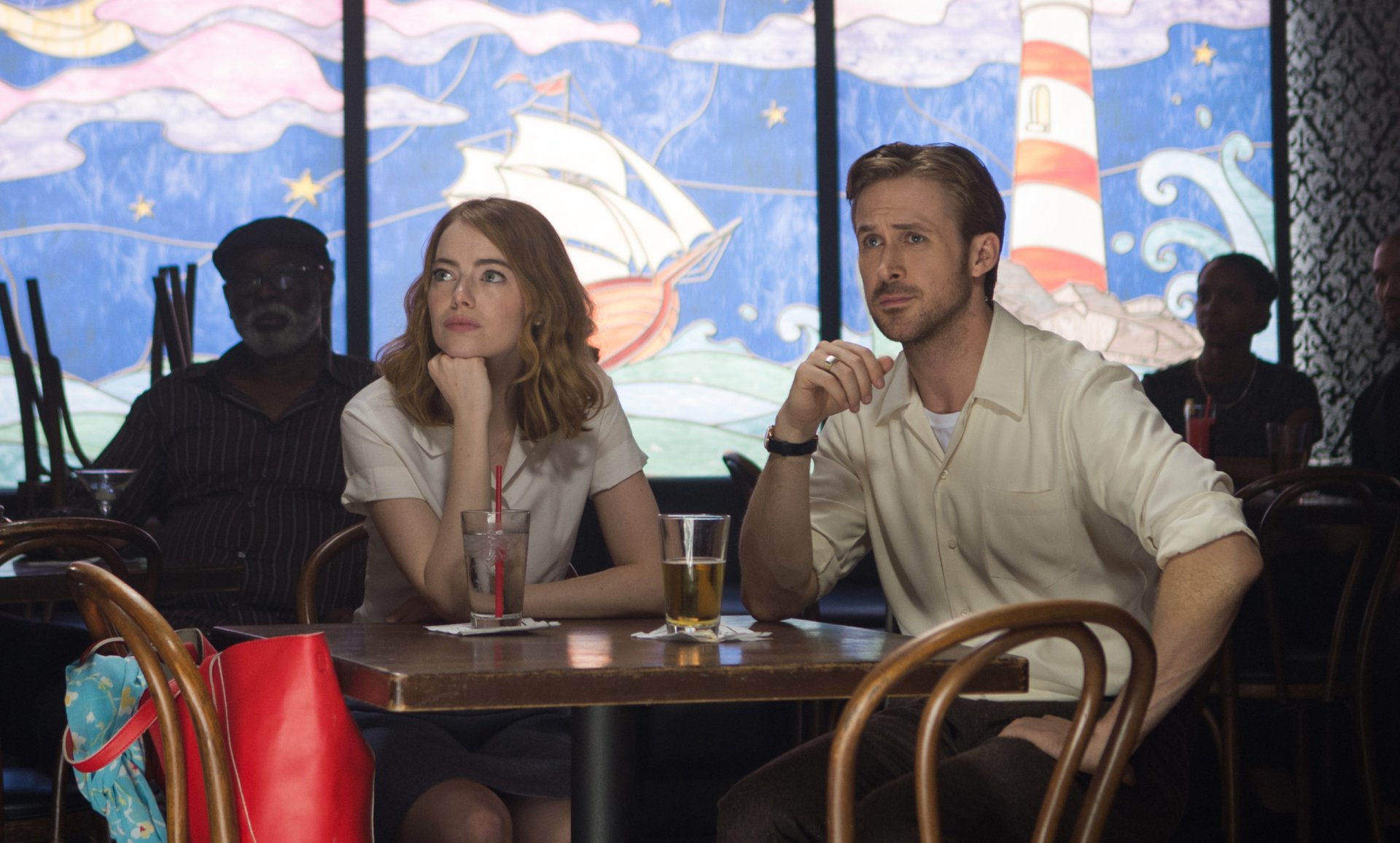 HD desktop wallpaper of Emma Stone and Ryan Gosling from the movie La La Land, sitting at a table with colorful, nautical-themed artwork in the background.