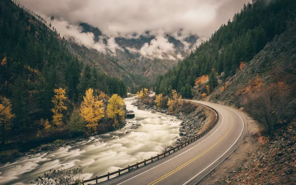 A scenic view of a winding road alongside a river, framed by forested mountains and autumn-colored trees, with foam gently rising on the water's surface.
