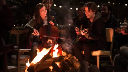 HD PC wallpaper from the movie If I Stay: a young woman plays cello and a young man plays guitar by a glowing campfire during an intimate outdoor night scene.