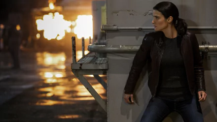 HD desktop wallpaper: woman in leather jacket crouches behind a metal column as an explosion lights a wet street — scene from Jack Reacher: Never Go Back.