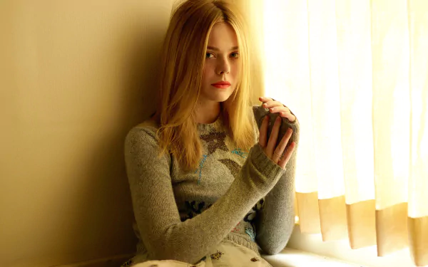 HD wallpaper of American actress Elle Fanning with green eyes and blonde hair, sitting by a softly lit window in a cozy, neutral-toned setting.
