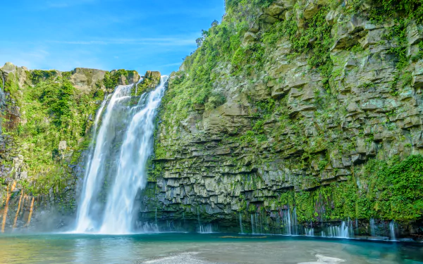 A stunning HD desktop wallpaper featuring a majestic waterfall cascading down a lush, green cliff into a serene pool below. The clear blue sky adds to the beauty of the natural scene.