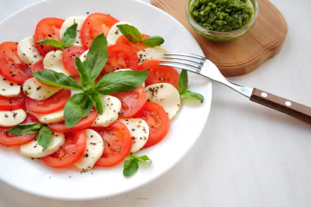 HD PC desktop wallpaper of a mozzarella and tomato salad: alternating slices of fresh mozzarella cheese and ripe tomatoes with basil, cracked pepper, a small bowl of pesto and a fork.