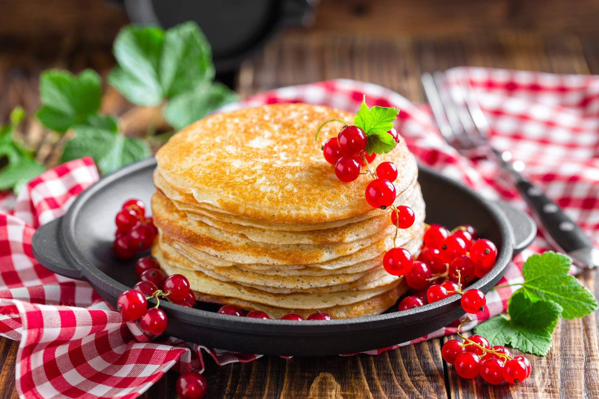 Still life of a stack of golden crêpes topped with red currants and green leaves on a skillet and red-checkered cloth — 5K Ultra HD PC wallpaper/background.