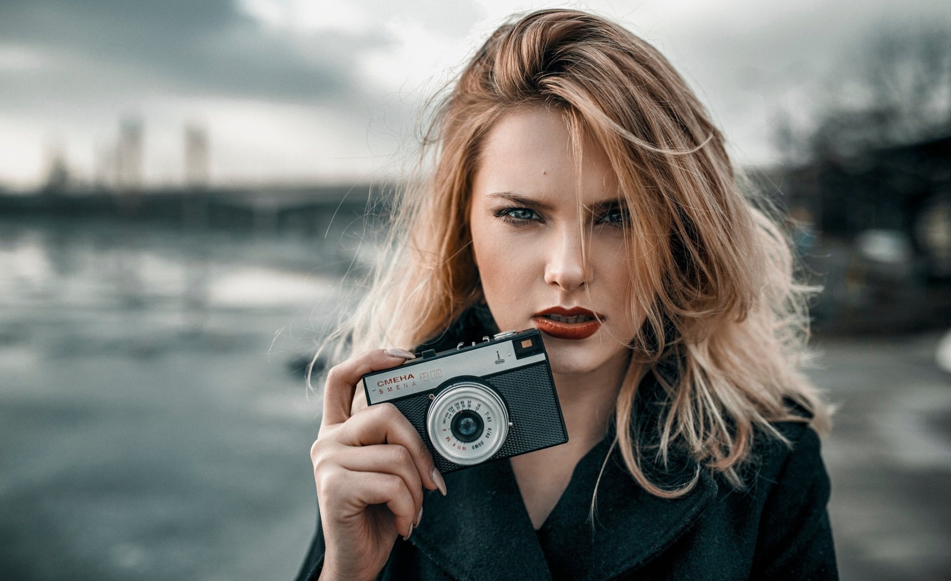 Blonde woman model with blue eyes and red lipstick holds a camera against a blurred cityscape, captured in sharp HD with a shallow depth of field for a striking desktop wallpaper.