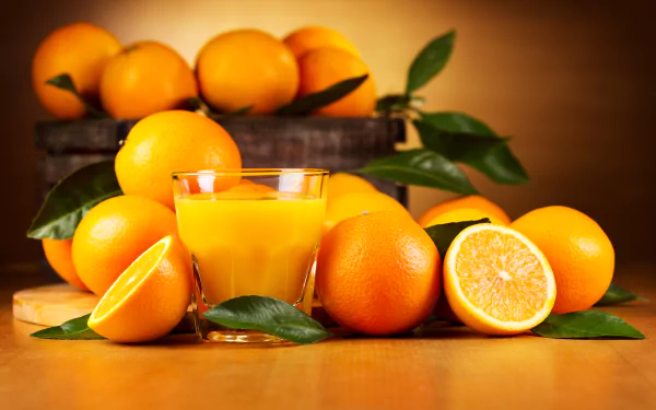 HD wallpaper featuring fresh oranges, green leaves, and a glass of orange juice against a warm background. The vibrant colors highlight the refreshing nature of the fruit and drink.