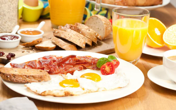 HD desktop wallpaper featuring a still life breakfast with fried eggs, crispy bacon, sliced bread, fresh orange juice, and coffee on a wooden table.