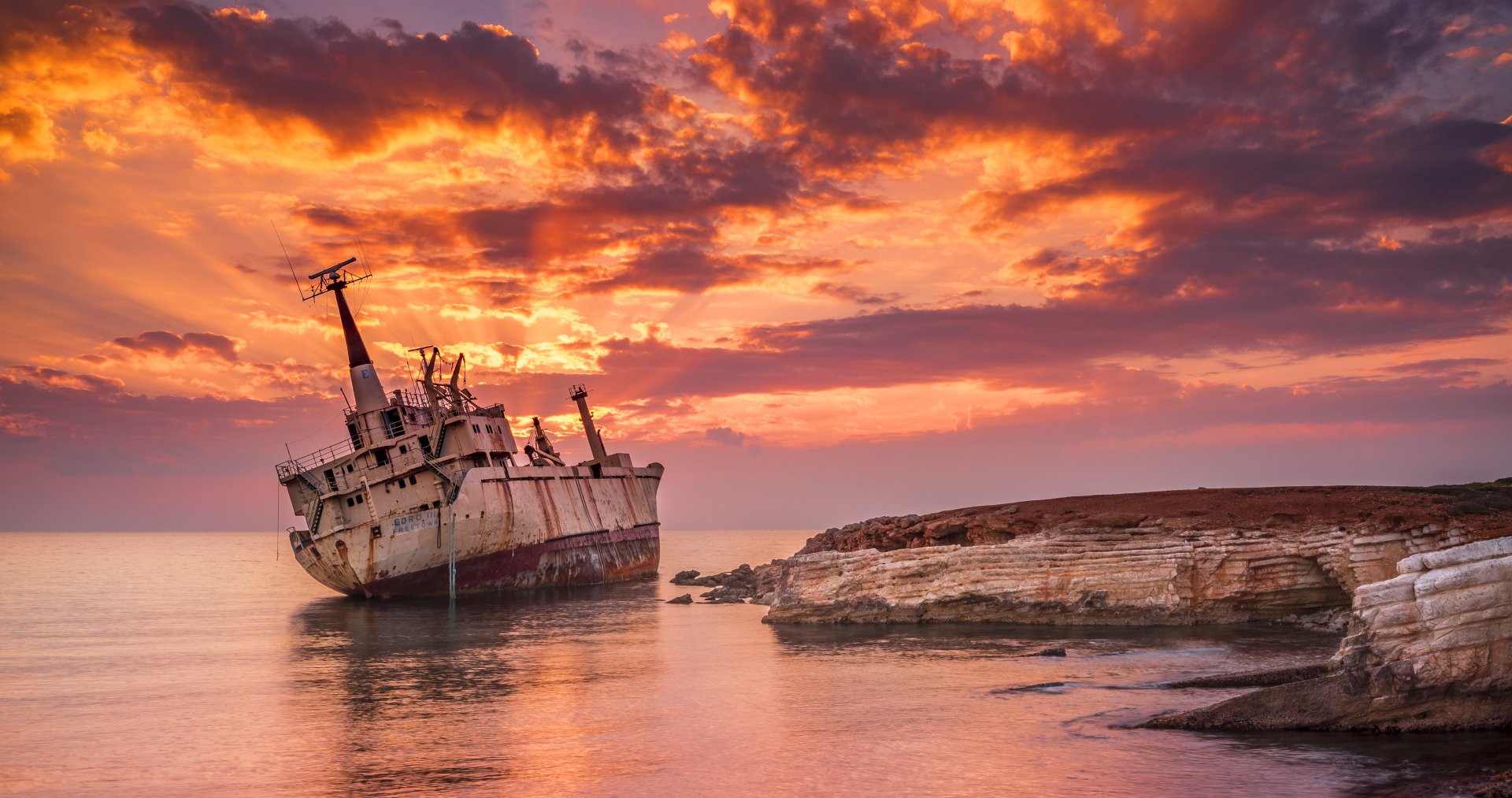 EDRO III cargo shipwreck rests near a cliff at sunset, with vibrant clouds reflecting on calm water in this 4K Ultra HD desktop wallpaper.
