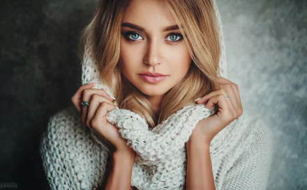 HD desktop wallpaper featuring a close-up of a blonde woman with blue eyes, wearing a knitted scarf. The background is textured, highlighting her striking gaze and serene expression.