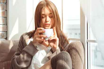 A woman with long hair, wearing a cozy sweater, sits by the window holding a cup, enjoying the sunlight. This HD desktop wallpaper and background exudes a calm and serene atmosphere.