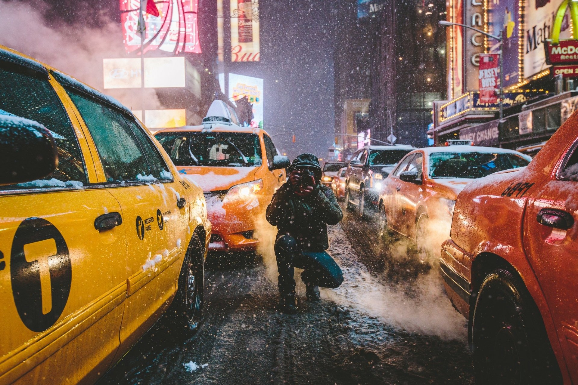 A winter night in New York City, featuring taxis lined up on a snowy street. The image captures intense snowfall and bright city lights, creating a classic urban scene in HD. A man is kneeling between the taxis.