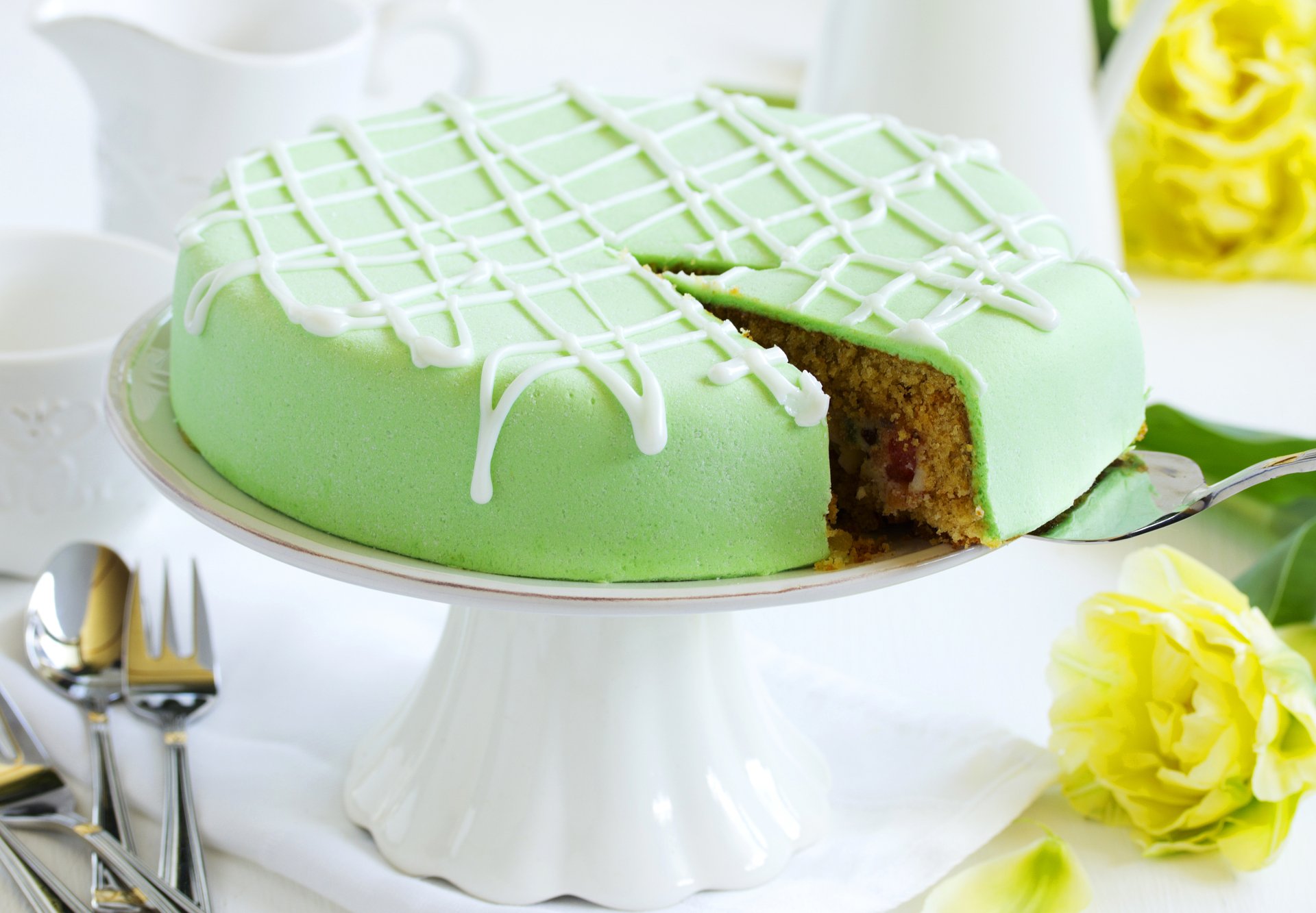 4K Ultra HD PC desktop wallpaper: green-iced pastry cake on a white pedestal with a slice removed, food styling with utensils and yellow flowers on a bright table.