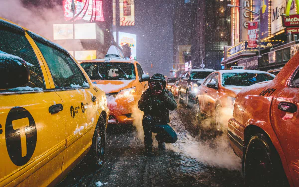 A winter night in New York City, featuring taxis lined up on a snowy street. The image captures intense snowfall and bright city lights, creating a classic urban scene in HD. A man is kneeling between the taxis.
