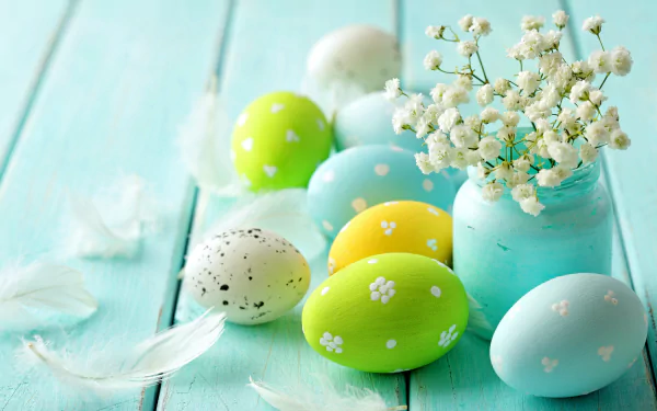 Colorful Easter eggs and white flowers in a jar on a light blue wooden surface, accompanied by delicate white feathers. HD desktop wallpaper and background for the Easter holiday.