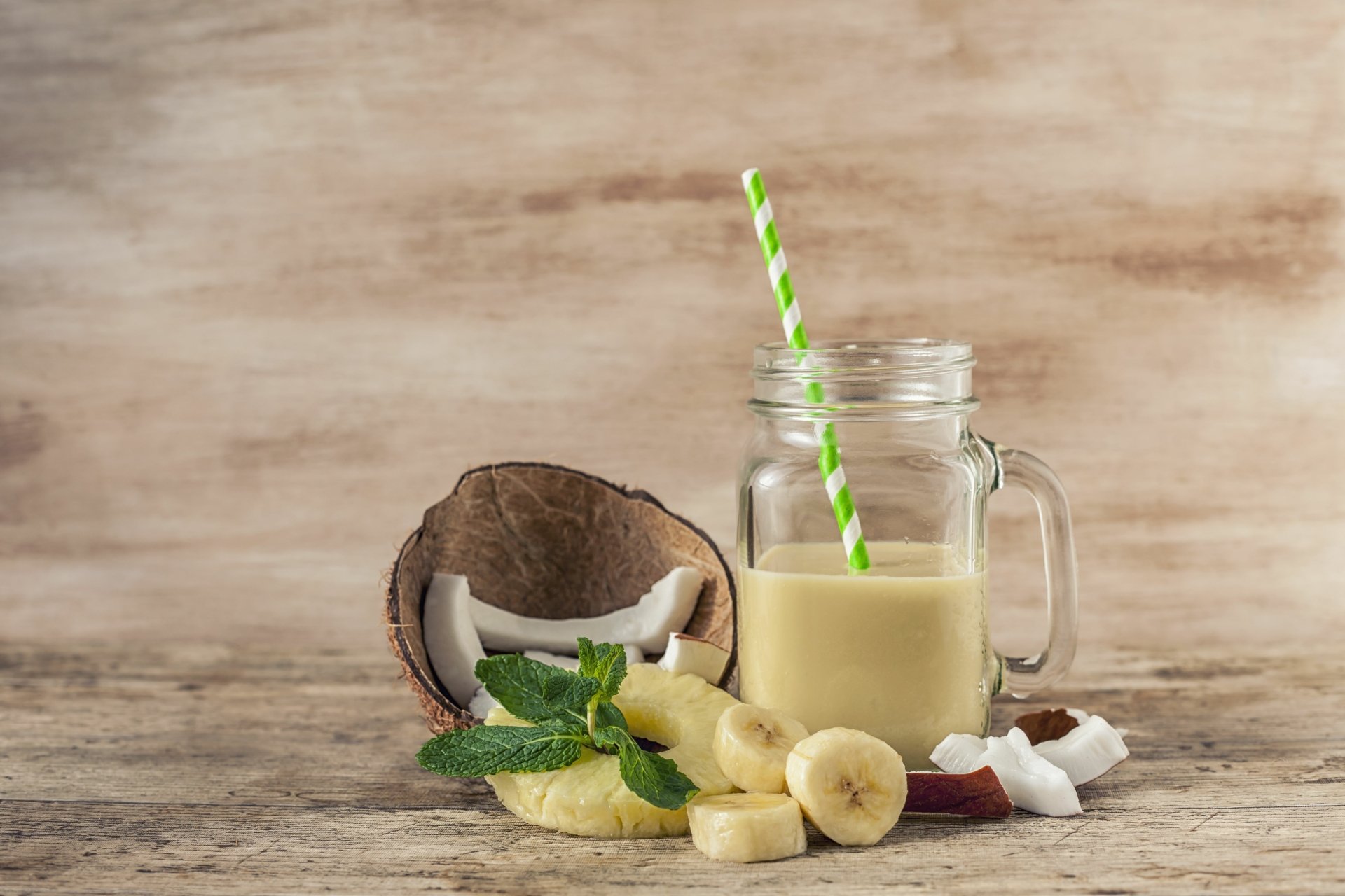4K Ultra HD wallpaper featuring a creamy coconut smoothie in a glass jar with a green-striped straw, sliced banana, coconut halves, and fresh mint on a rustic wooden surface.