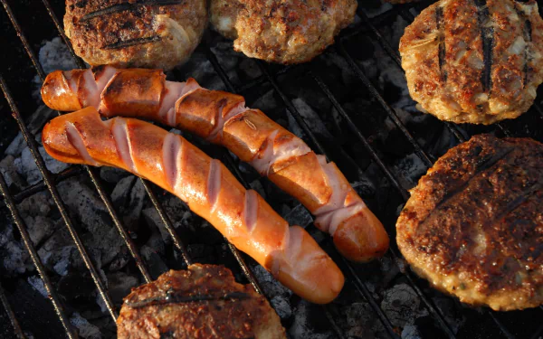Close-up HD desktop wallpaper of sizzling sausages and burger patties cooking on a barbecue grill.