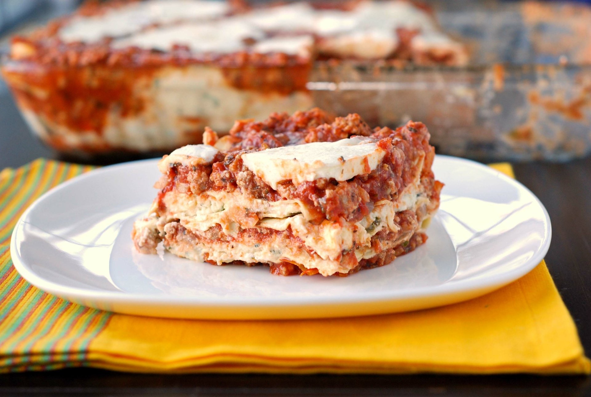 A close-up HD desktop wallpaper of a slice of layered lasagna served on a white plate with the remaining dish in the background, highlighting a hearty meal.