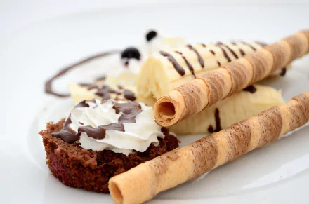 4K Ultra HD PC desktop wallpaper showing a close-up dessert: banana slice, whipped cream-topped biscuit, chocolate drizzle and two rolled wafer sticks on a white plate.