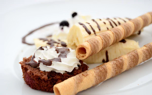 4K Ultra HD PC desktop wallpaper showing a close-up dessert: banana slice, whipped cream-topped biscuit, chocolate drizzle and two rolled wafer sticks on a white plate.