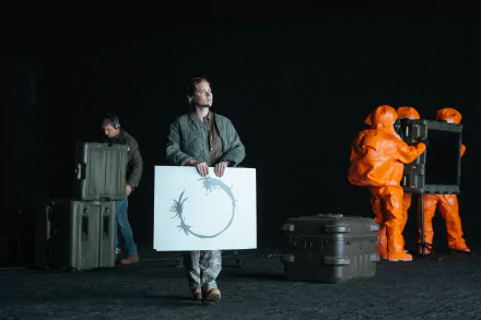 Scene from the movie Arrival featuring Amy Adams holding a circular symbol, with two people in orange hazmat suits and another person handling equipment in the background.