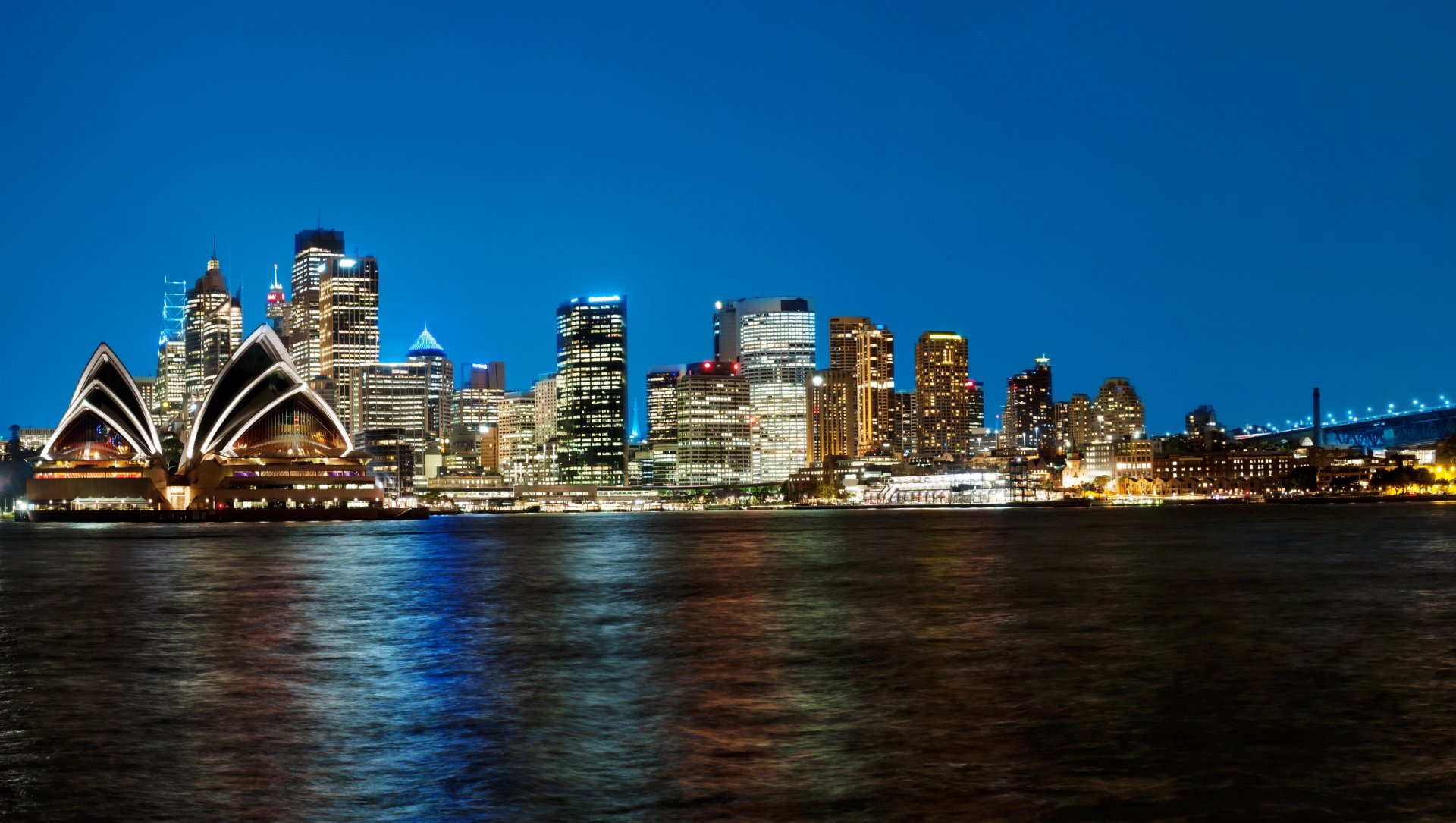 HD desktop wallpaper showcasing the Sydney Opera House and city skyscrapers illuminated at night, reflecting on the water in Australia.