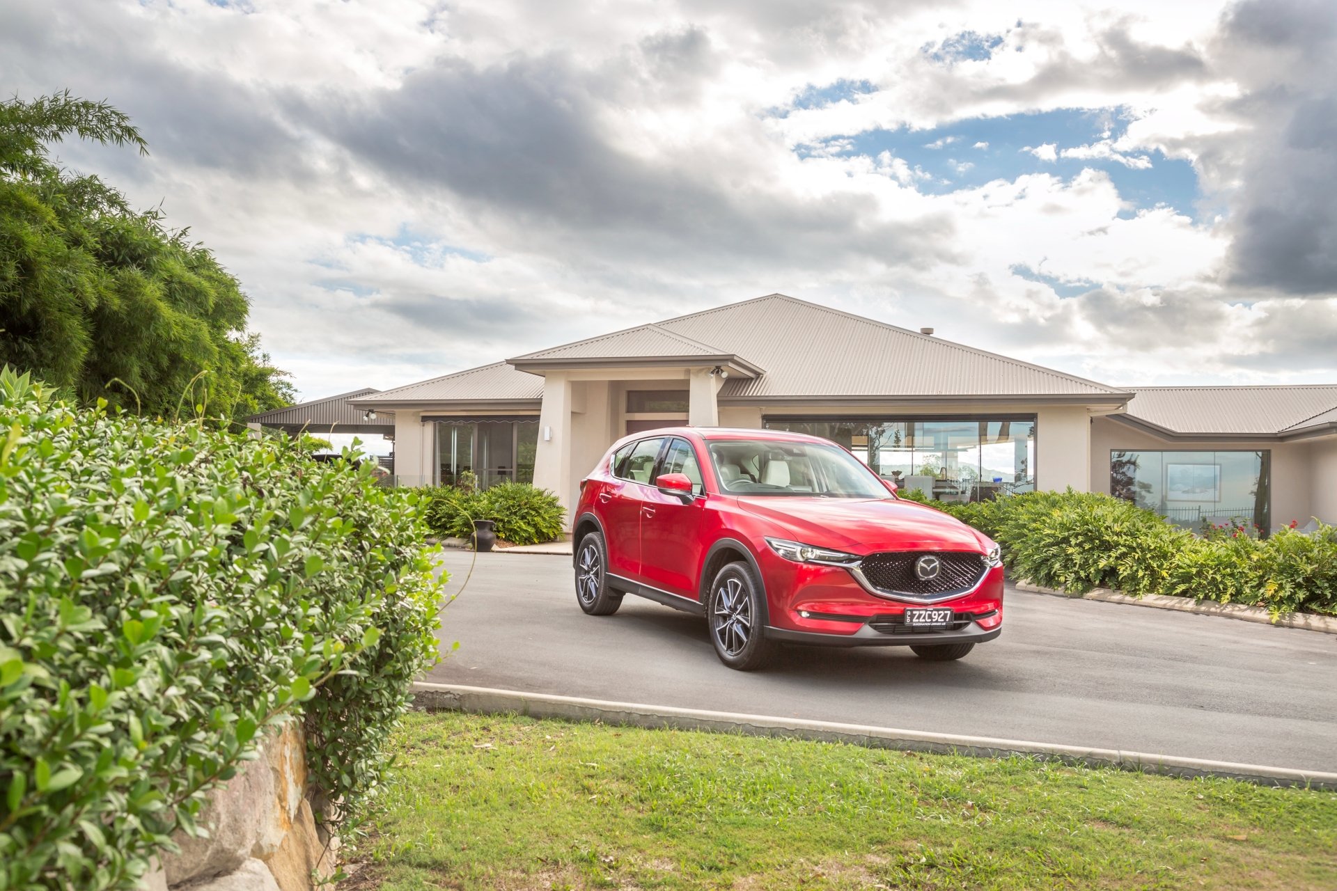 Red Mazda CX-5 SUV car parked before a modern house under dramatic clouds, 4K Ultra HD PC desktop wallpaper background.