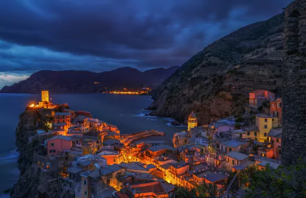 Nighttime view of Vernazza, a picturesque coastal village in Cinque Terre, Italy, with brightly lit houses perched on cliffs along the rugged coastline.