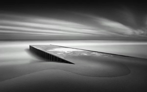 Black and white HD desktop wallpaper of a barren beach landscape with smooth sand and a partially submerged pier extending into calm water.