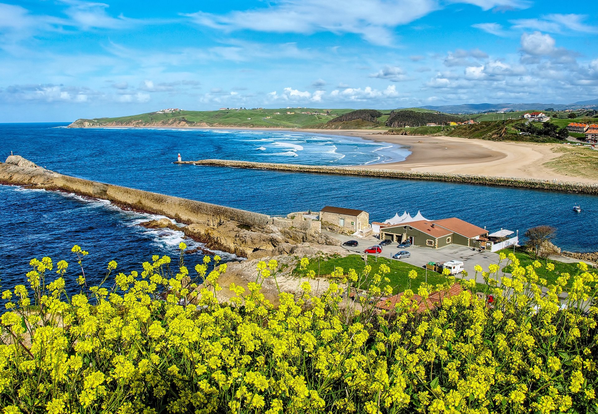 HD desktop wallpaper of Cantabria’s coastline in Spain, featuring a sandy beach, yellow rapeseed flowers, a house by the sea, and the vast ocean under a blue sky.