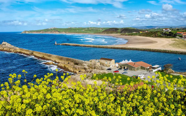 HD desktop wallpaper of Cantabria’s coastline in Spain, featuring a sandy beach, yellow rapeseed flowers, a house by the sea, and the vast ocean under a blue sky.