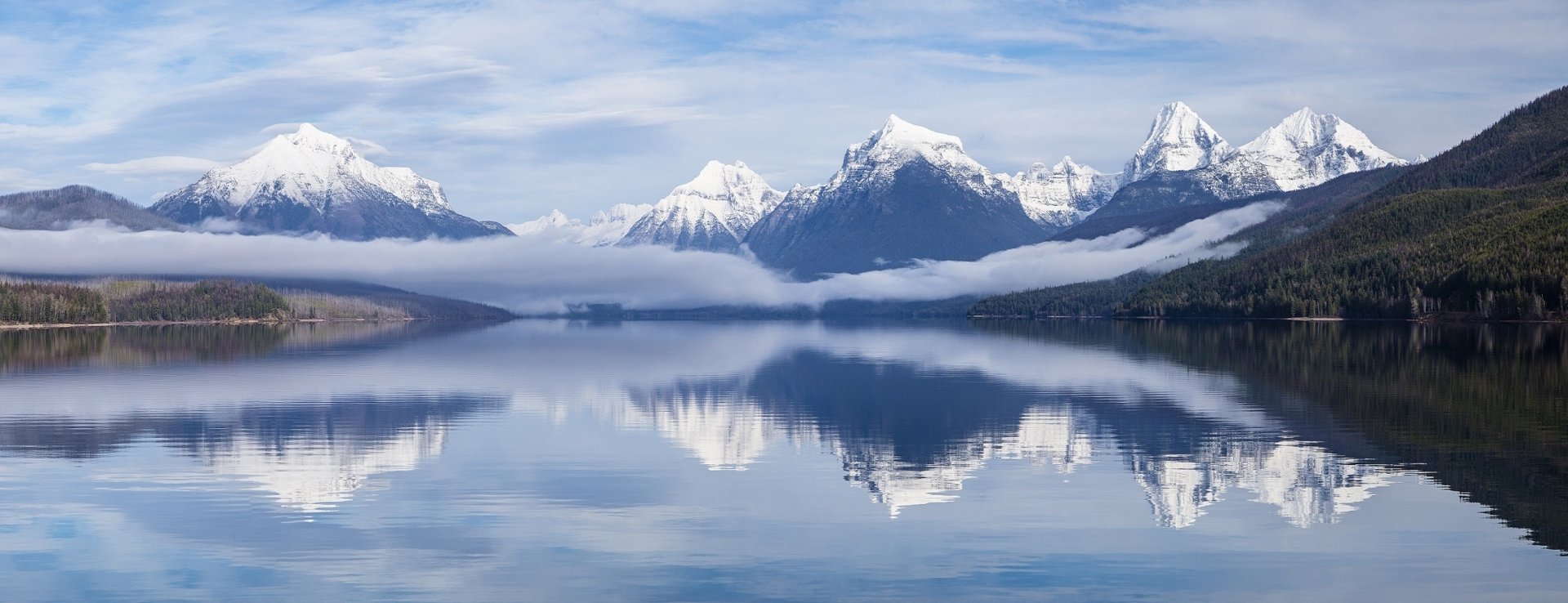 Panoramic view of Lake McDonald in Glacier National Park, Montana, USA. Snow-capped mountains and fog are reflected in the calm lake, creating a serene HD wallpaper and background.