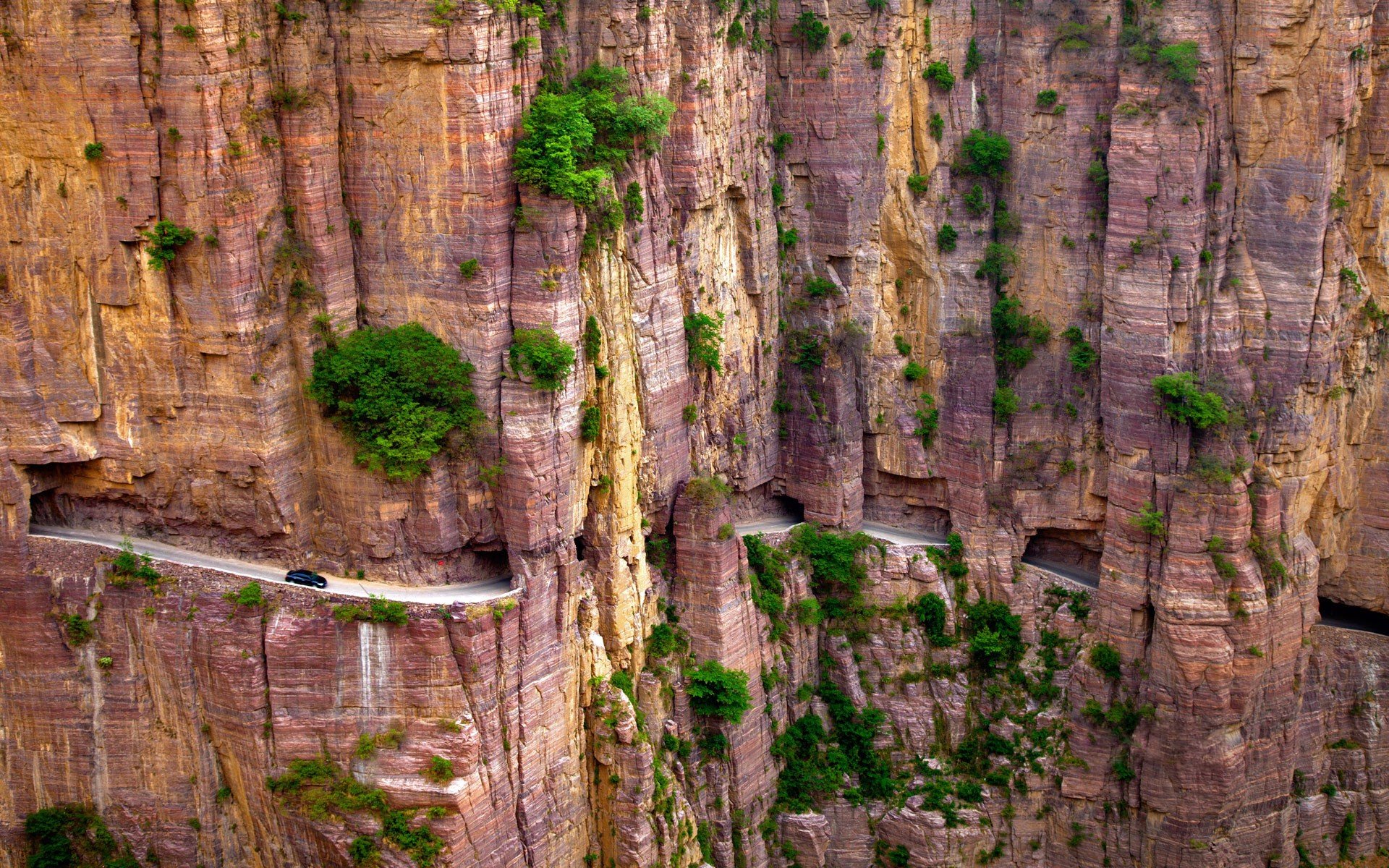 High-definition desktop wallpaper featuring a man-made tunnel road carved into a dramatic, vertical mountain landscape in China, with patches of greenery dotting the rugged rock face.