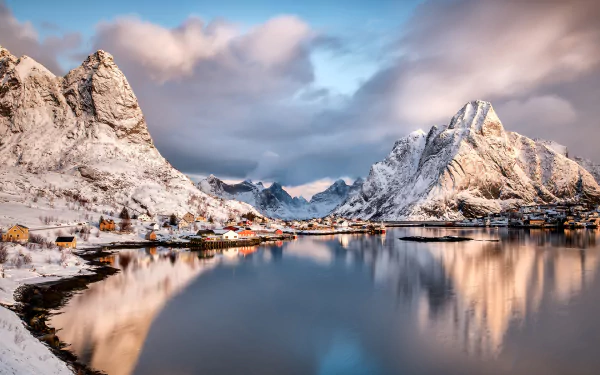 Snow-covered mountains frame the serene village of Reine in Lofoten, Norway, reflected in calm waters under a soft, cloudy sky.