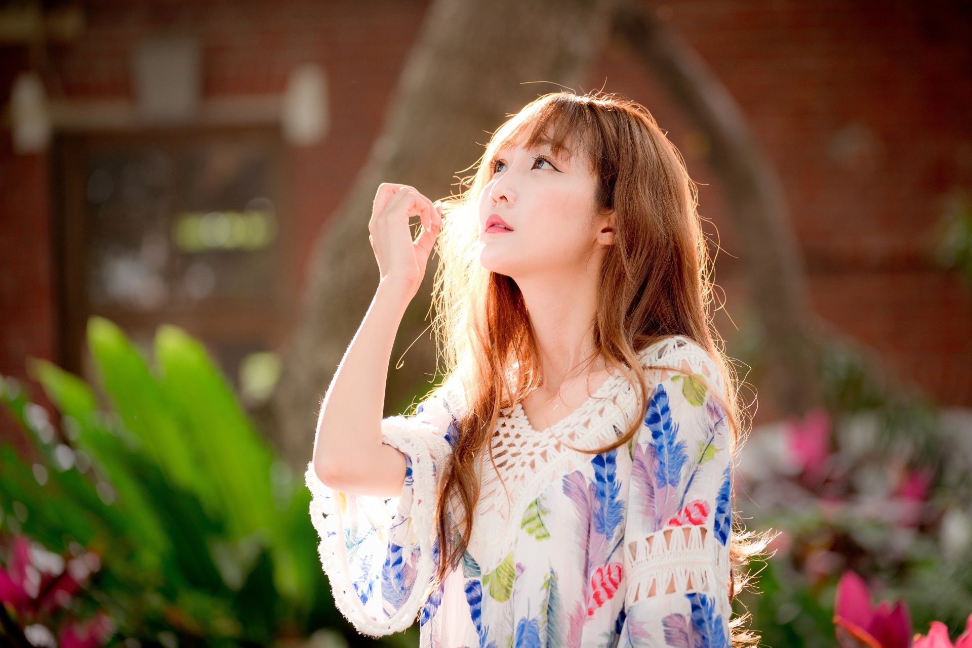 HD desktop wallpaper featuring a serene mood, showcasing a red-haired, blue-eyed Asian woman. The depth of field highlights the model as she gazes thoughtfully, surrounded by lush greenery and soft lighting.