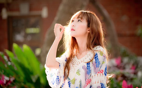 HD desktop wallpaper featuring a serene mood, showcasing a red-haired, blue-eyed Asian woman. The depth of field highlights the model as she gazes thoughtfully, surrounded by lush greenery and soft lighting.