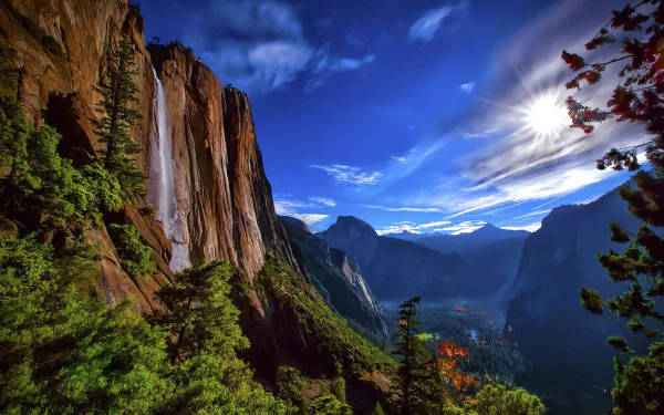 HD desktop wallpaper of Yosemite National Park featuring a stunning landscape with a waterfall cascading down a rugged cliff, a lush forest, a serene valley, and majestic mountains under a vibrant sky.