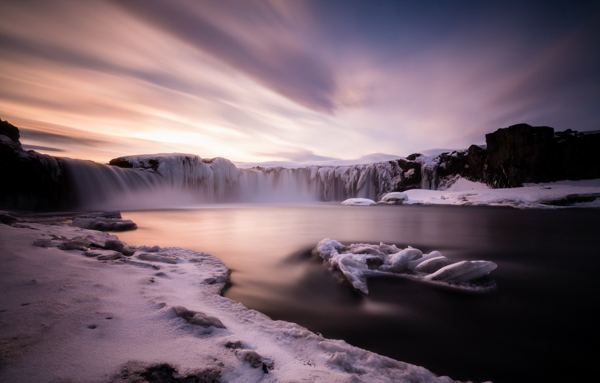 A stunning HD desktop wallpaper of Goðafoss waterfall in Iceland, featuring cascading water, snowy banks, a dramatic sky, and soft cloud formations over tranquil water.