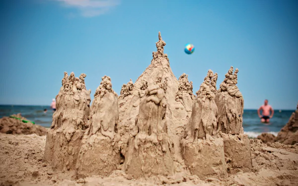 Artistic sandcastle on a beach, detailed sculpted castle with ocean and a blurred beach ball and swimmer — 4K Ultra HD PC desktop wallpaper/background.