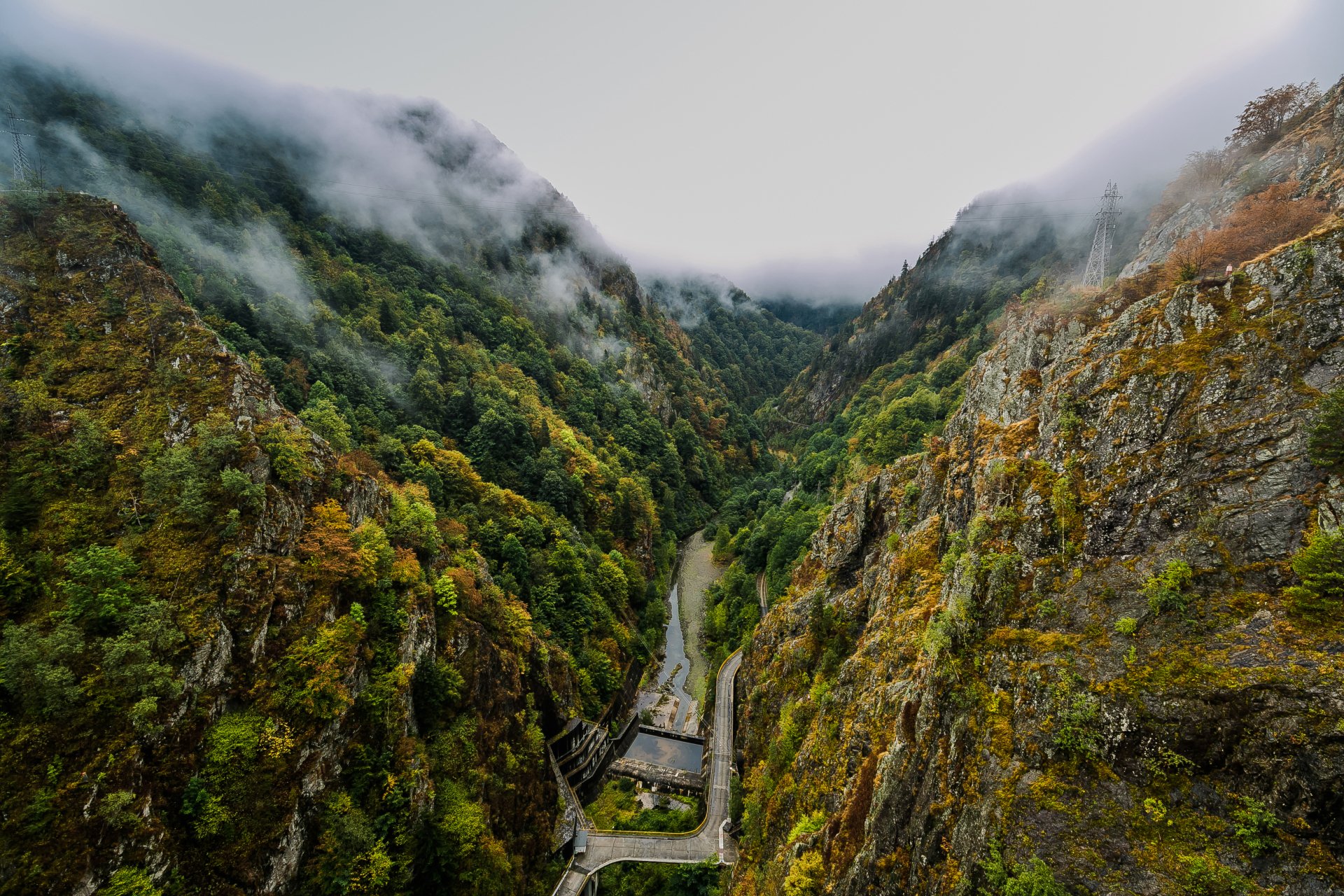 Romania mountain forest landscape, aerial photography of a narrow green valley with winding road and small riverside building, dramatic cliffs and mist — 2K Quad HD PC desktop wallpaper/background.