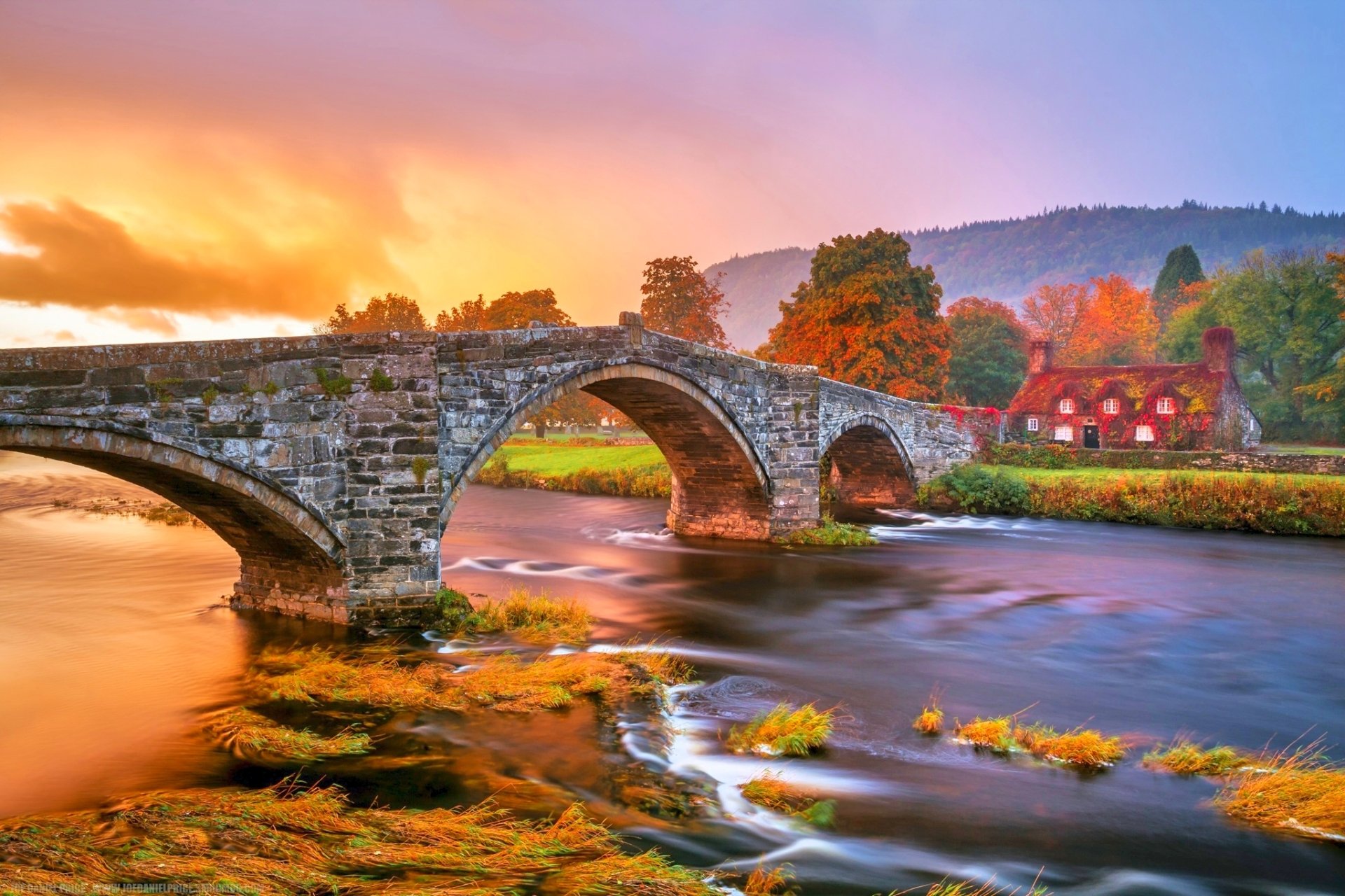 HD PC desktop wallpaper: autumn river flowing beneath a stone man-made arch bridge, ivy-draped banks and colorful fall trees beside a cottage house.