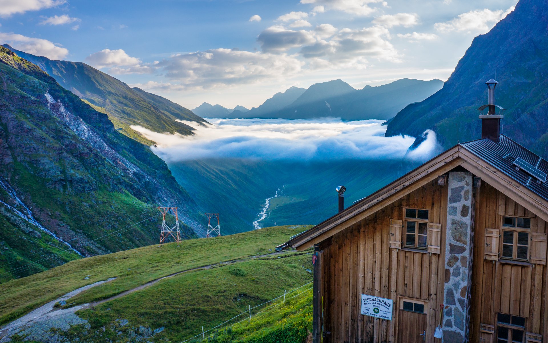 A breathtaking HD desktop wallpaper of an Austrian mountain landscape with clouds nestled in the valley and a rustic wooden cabin in the foreground.