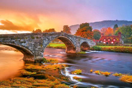 HD PC desktop wallpaper: autumn river flowing beneath a stone man-made arch bridge, ivy-draped banks and colorful fall trees beside a cottage house.