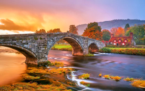 HD PC desktop wallpaper: autumn river flowing beneath a stone man-made arch bridge, ivy-draped banks and colorful fall trees beside a cottage house.
