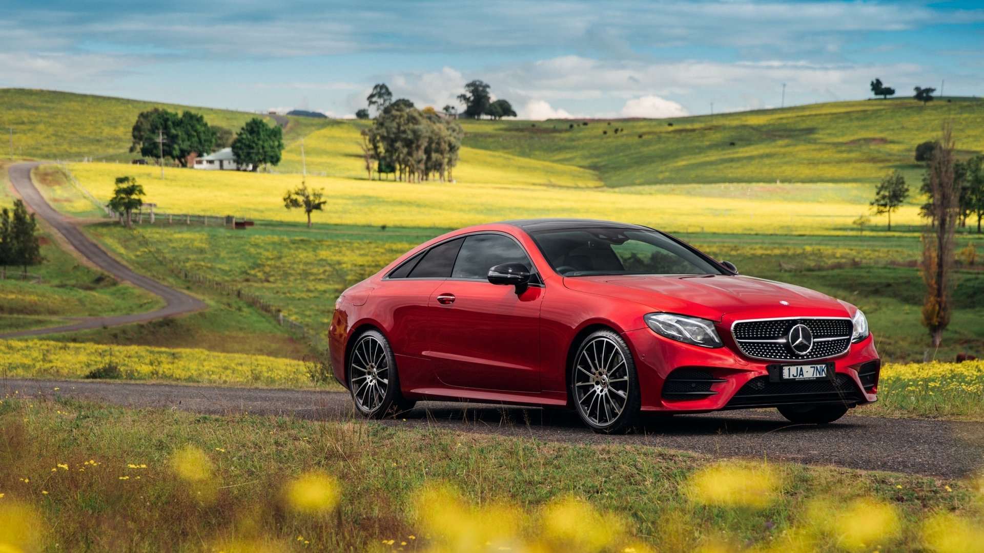 A bright red Mercedes-Benz E-Class car is parked on a countryside road surrounded by lush green fields with yellow flowers, under a partly cloudy sky. HD desktop wallpaper and background.