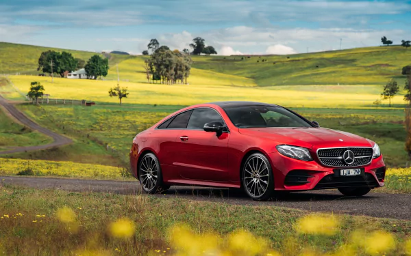 A bright red Mercedes-Benz E-Class car is parked on a countryside road surrounded by lush green fields with yellow flowers, under a partly cloudy sky. HD desktop wallpaper and background.