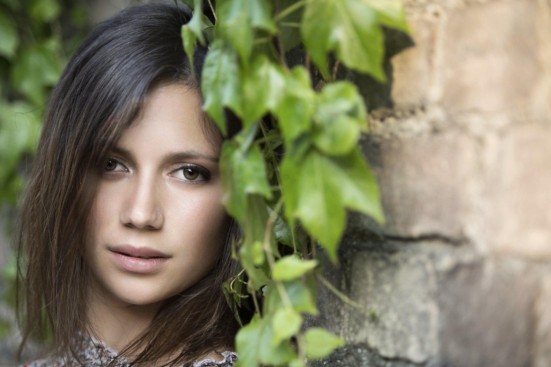 HD desktop wallpaper featuring a brunette model with brown eyes, posing next to an old brick wall adorned with green ivy.